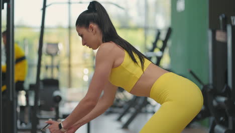 A-young-Hispanic-woman-in-a-yellow-tracksuit-performs-an-exercise-in-a-crossover-pulls-a-steel-rope-from-below-to-train-her-back.-A-woman-trains-her-back-in-a-gymnasium